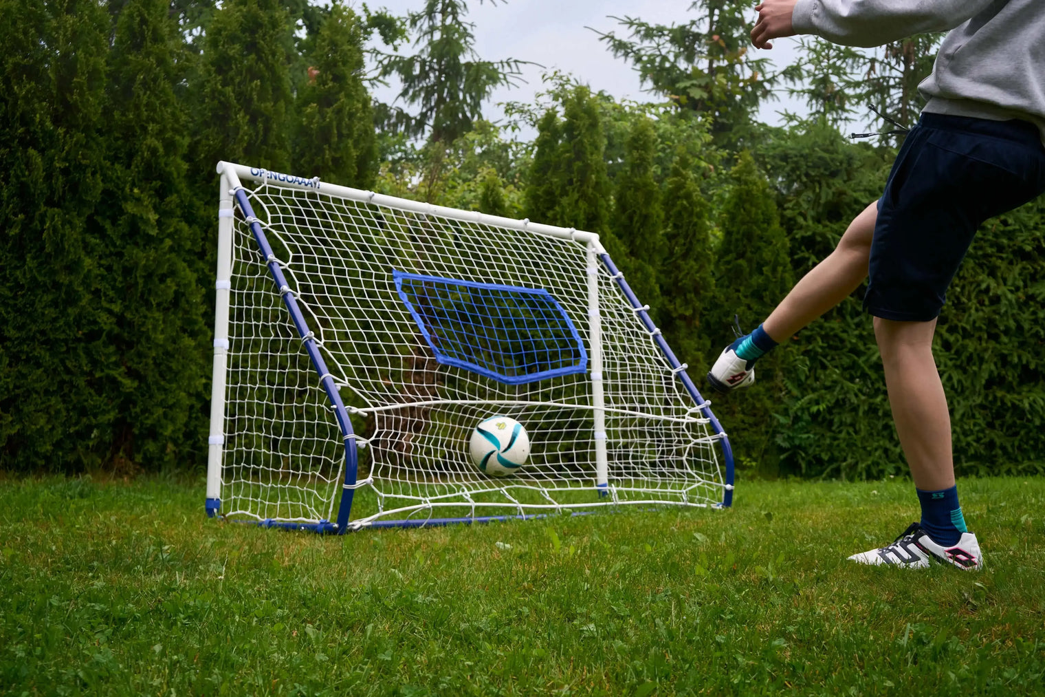 A person kicking a soccer ball toward a compact football rebounder goal trainer in a backyard setting.