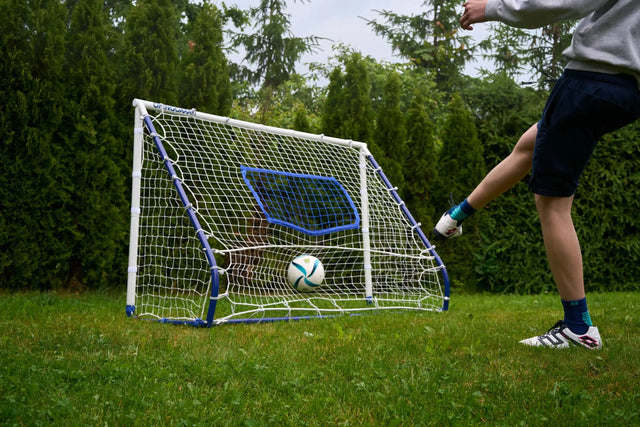 A person kicking a soccer ball toward a compact football rebounder goal trainer in a backyard setting.