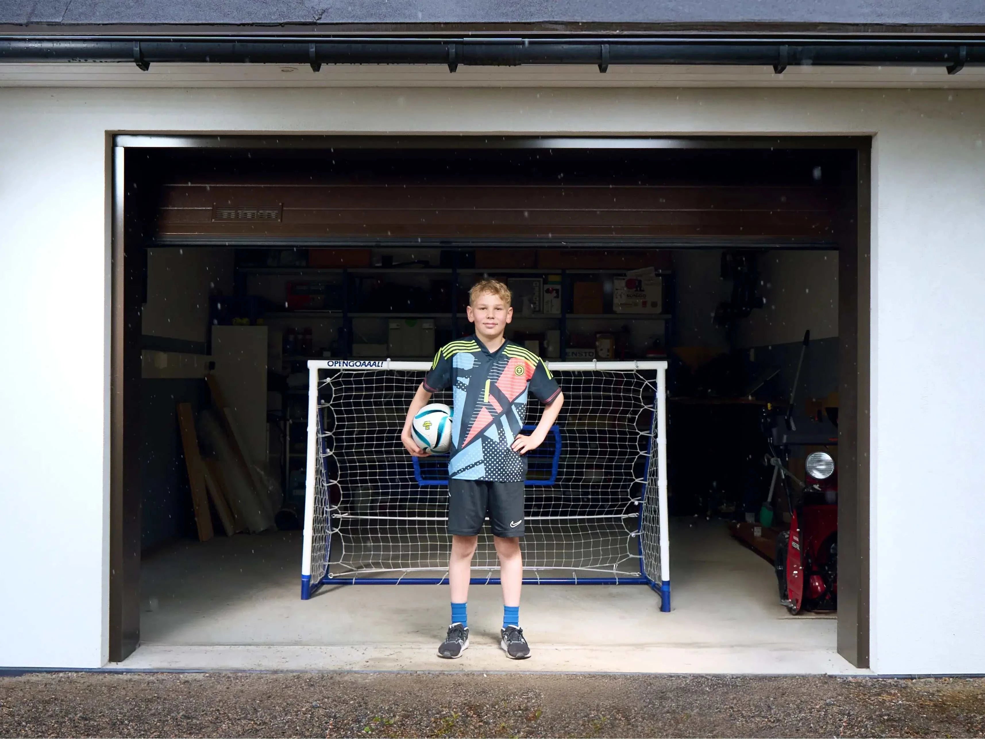 Young boy in soccer gear with a football in front of a goal in a garage setting, ready for practice.
