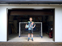 Young boy in soccer gear with a football in front of a goal in a garage setting, ready for practice.