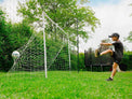 Person kicking a soccer ball towards a goal on a grassy field with trees in the background