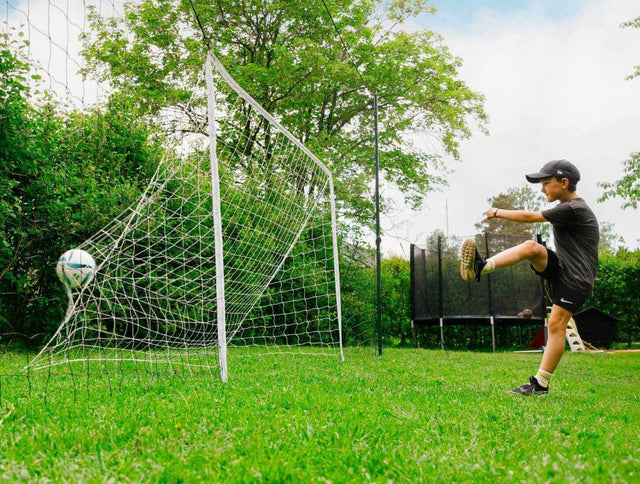 Person kicking a soccer ball towards a goal on a grassy field with trees in the background