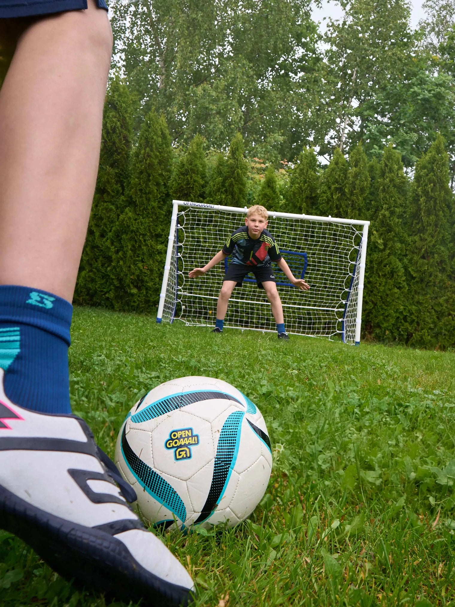 Child practicing soccer with a football rebounder goal trainer in a backyard, aiming at a small goal.