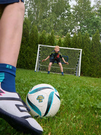 Child practicing soccer with a football rebounder goal trainer in a backyard, aiming at a small goal.