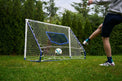 A person kicking a soccer ball toward a compact football rebounder goal trainer in a backyard setting.