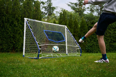 A person kicking a soccer ball toward a compact football rebounder goal trainer in a backyard setting.